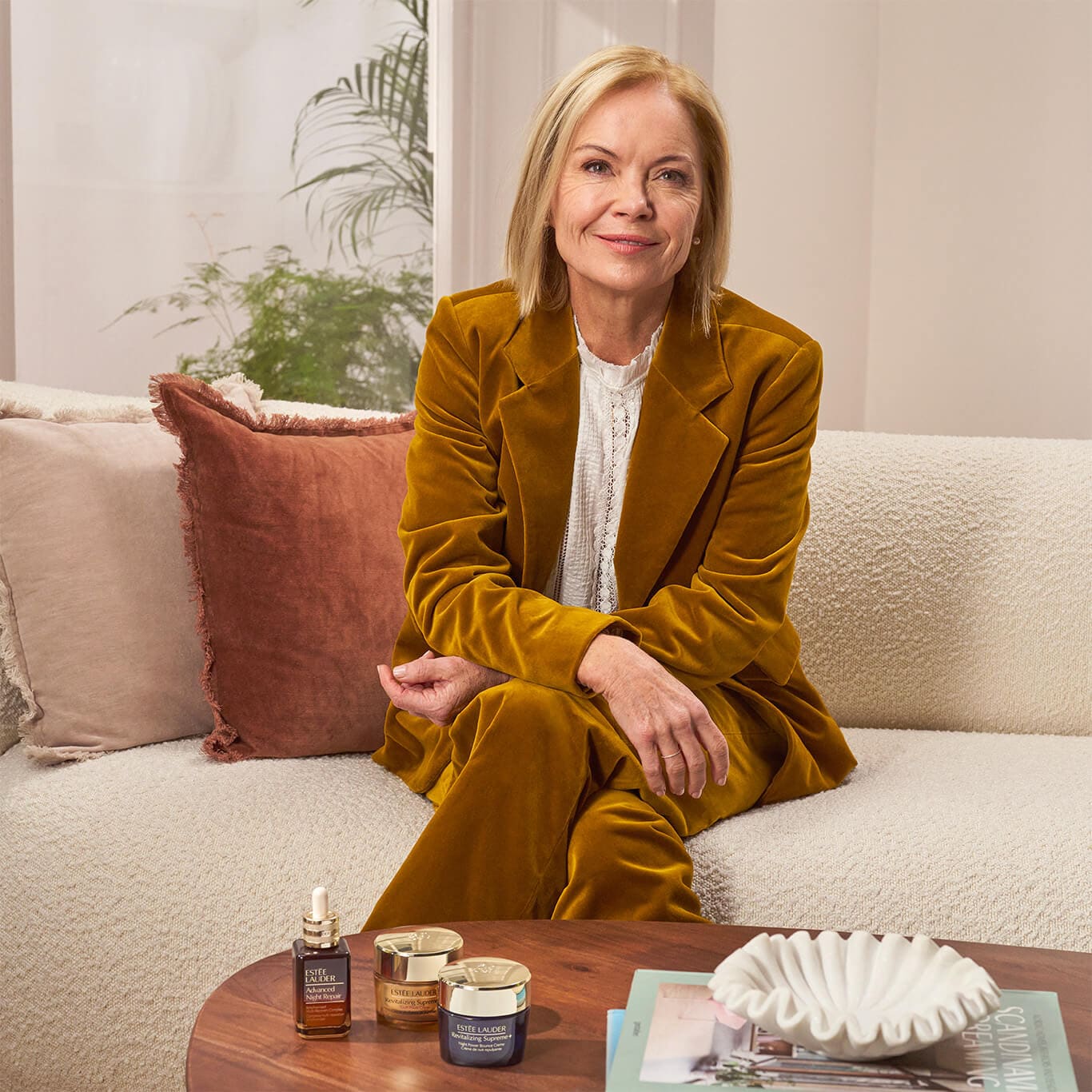 Mariella Frostup sitting on a sofa with our best selling skincare products sitting on a table in front of her